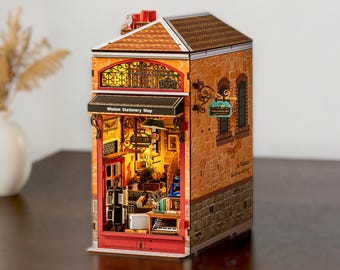 May include: A miniature model of a Wisdom Stationery Shop with a stone-textured exterior, a red door frame, and a lit interior. The building features a brown roof, a hanging sign, and detailed shelves filled with tiny books and office supplies.