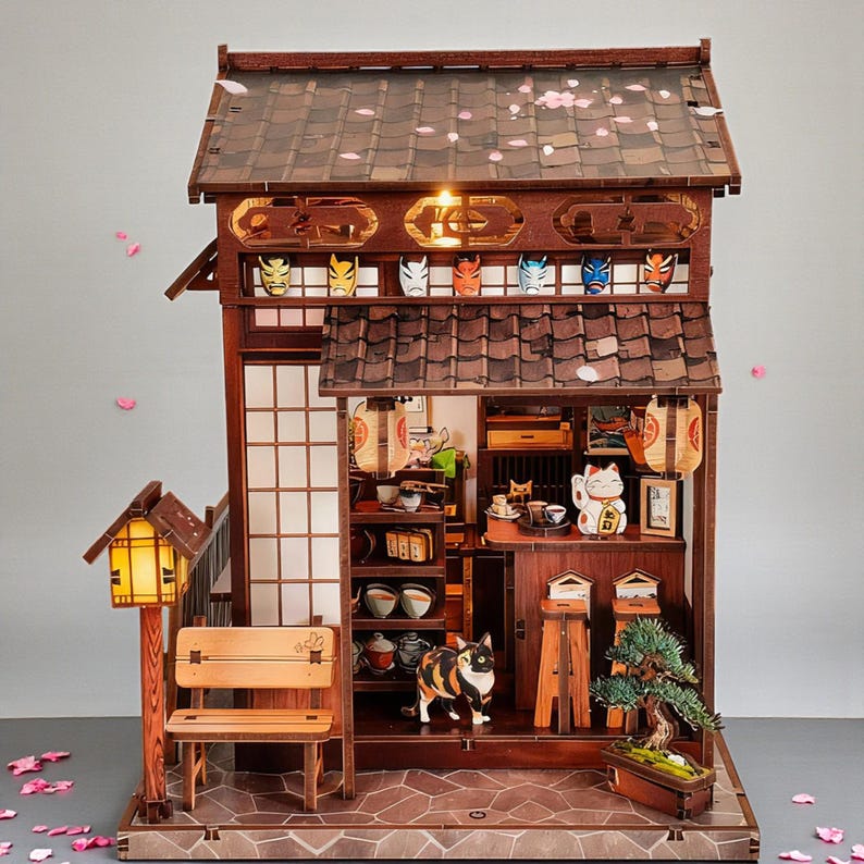 May include: A detailed wooden miniature Japanese house with a tiled roof, paper screen doors, and a small bench. The interior features miniature furniture, a cat figurine, and decorative masks. A bonsai tree and paper lanterns add to the scene.