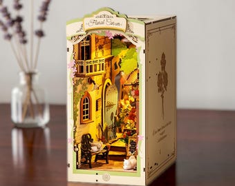 May include: A wooden book nook kit titled Floral Corner featuring a miniature street scene with a bench, a cat, and warm interior lighting. The structure includes a balcony, a green door, and floral details, with text printed on the side panel.
