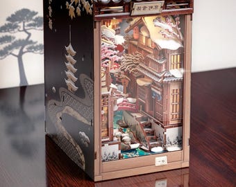 May include: A 3D wooden book nook diorama featuring a miniature Japanese street scene with snow-covered rooftops, a red bridge, and cherry blossom trees. The black exterior side panel displays a pagoda illustration and Japanese text characters.