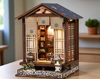 May include: A miniature wooden Japanese house model with intricate details. The model features a dark brown frame, a tiled roof, and paper screen windows. The interior is lit, revealing a small library and miniature figures. The base is decorated with stones and miniature plants.