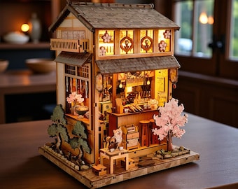 May include: A detailed miniature wooden Japanese restaurant model, featuring a bar, stools, and hanging lanterns. The model has a tiled roof, decorative windows, and a small cherry blossom tree. The scene is illuminated with warm lighting, creating a cosy atmosphere.