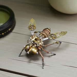Puede incluir: Una detallada escultura de insecto de estilo steampunk. El insecto metálico presenta componentes de latón, cobre y plata, incluyendo engranajes, tubos y patas articuladas. Las alas tienen un delicado diseño de filigrana. La escultura está sobre una superficie de madera clara.
