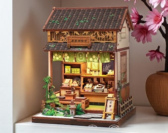 Peut inclure: Maquette miniature d'une boutique japonaise avec un toit en tuiles marron, des accents de bambou et des auvents en tissu vert. L'intérieur est éclairé, révélant des étagères avec des produits miniatures. Un petit panneau est devant, et un vase de fleurs roses à droite.