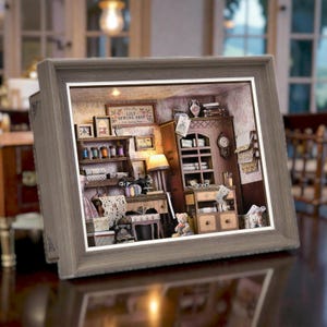 Peut inclure: Un diorama miniature d'un atelier de couture encadré dans un cadre en bois gris. La scène comprend une armoire en bois, une machine à coudre, des étagères avec des bobines de fil, un petit ours en peluche et une enseigne Lily Sewing Shop.