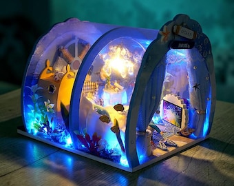 May include: A miniature dollhouse kit with an ocean theme, featuring blue arched frames, interior LED lighting, and small furniture. The structure includes a sign reading OCEAN HOME, decorative fish cutouts, a small television screen, and fabric curtains.
