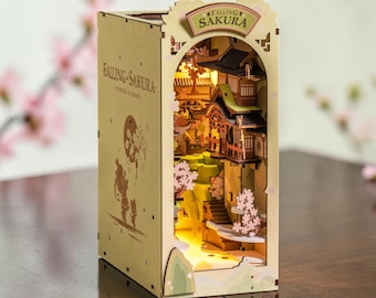 May include: A light-colored wooden book nook insert titled Falling Sakura, featuring an illuminated interior with layered architectural structures, miniature staircases, and pink cherry blossom accents. The exterior side panel displays the text Falling Sakura.