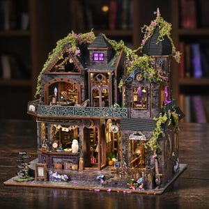 May include: A detailed miniature dollhouse with a witchy theme. The multi-story structure features a dark gray brick facade, ornate windows, and a balcony. The house is adorned with faux greenery and purple lighting, with the words "Witch's Lair" visible.