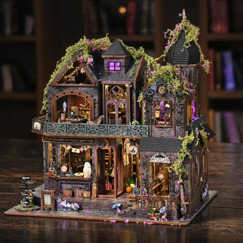 May include: A detailed miniature dollhouse with a witchy theme. The multi-story structure features a dark gray brick facade, ornate windows, and a balcony. The house is adorned with faux greenery and purple lighting, with the words "Witch's Lair" visible.