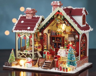 May include: A miniature Christmas-themed dollhouse featuring a red roof with white snow accents, a brick chimney, and an open interior. The scene includes a snowman, two small Christmas trees, wrapped gifts, a porch, and interior lighting.