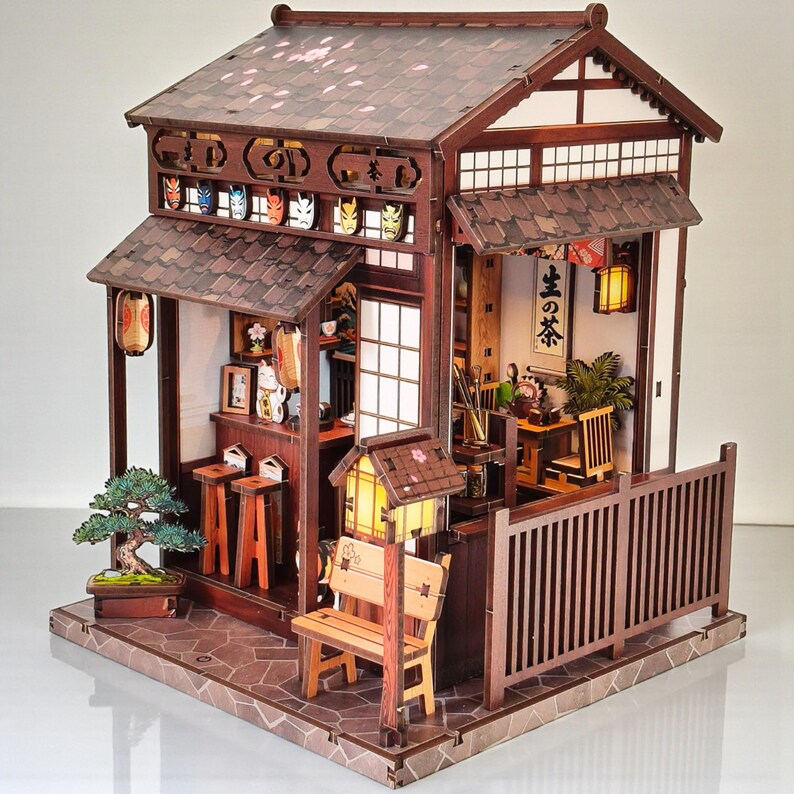 May include: A detailed wooden miniature Japanese tea house with a brown tiled roof, paper lanterns, and a small bench. The facade features decorative masks and the interior includes miniature furniture and plants.