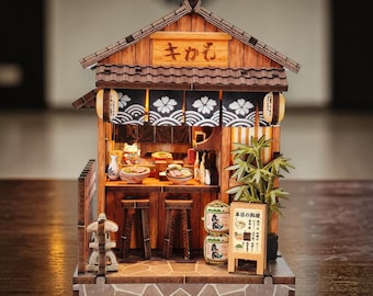 May include: A miniature wooden Japanese ramen shop model with a detailed exterior, including a sign with Japanese characters, lanterns, and a small menu board. The interior features a counter, stools, and miniature food items.