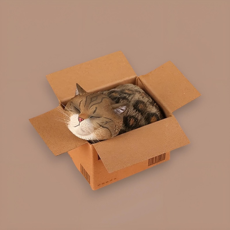 May include: A brown and white tabby cat figurine sleeping inside a cardboard box. The box is open and the cat is curled up inside.