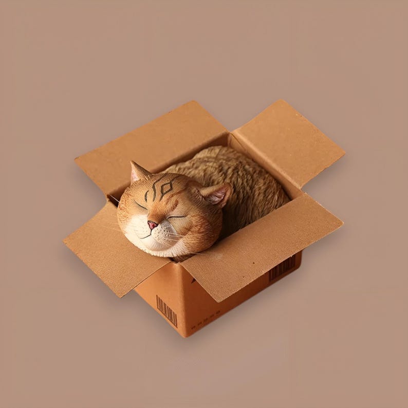 May include: A brown and white cat figurine sleeping inside a cardboard box. The cat is curled up in a ball with its eyes closed.