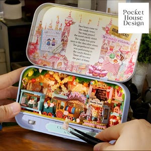 May include: A miniature dollhouse scene inside a tin box. The scene is a candy town with a train, a carousel, and a candy store. The box lid has a whimsical illustration of a candy town with the text "It's candy o'clock." and "Golden Ticket".