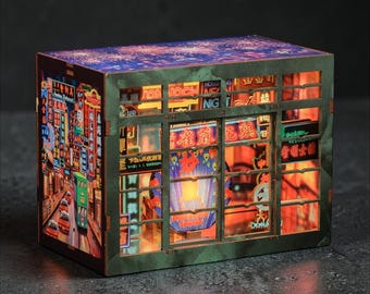 May include: A miniature, illuminated diorama depicting a bustling city street scene at night. The detailed model features colorful neon signs, buildings, and streetlights, creating a vibrant and immersive display. The diorama is housed in a rectangular frame.