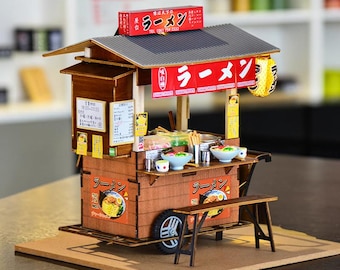 May include: A miniature wooden ramen food stall model featuring a dark grey corrugated roof, a red sign with Japanese text, a yellow paper lantern, and a small bench. The structure includes detailed menu boards, food bowls, and wheels.