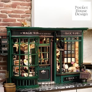 Könnte beinhalten: Ein Miniatur-Puppenhaus-Laden mit grüner Fassade und einem Schild mit der Aufschrift "Magic Wand Magical Wand Shop". Die Schaufenster des Ladens sind mit verschiedenen Gegenständen gefüllt, darunter Bücher, Zauberstäbe und andere magische Gegenstände.