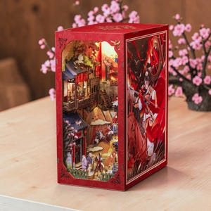 May include: A red, illuminated, laser-cut wooden book nook with a detailed miniature Asian village scene. The book nook has a detailed illustration on the side. The scene includes buildings, figures, and cherry blossom trees.