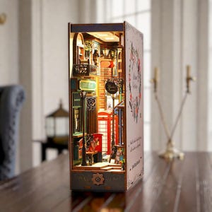 Könnte beinhalten: Ein hölzernes Buchnischen-Diorama mit einer Straßenszene im Miniaturformat, inklusive roter Telefonzelle, Postamt-Schild, Ziegelwänden und warmer Innenbeleuchtung. Die äußere Seitenwand zeigt eine florale Kranzgrafik und gedruckten Text.