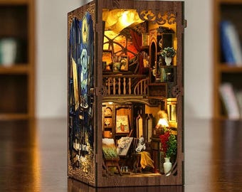 May include: A miniature wooden diorama of an artist's studio, illuminated from within. The multi-level structure features detailed furniture, artwork, and a spiral staircase. The exterior is a dark brown with intricate cut-out designs, and the interior is lit with a warm, inviting glow.