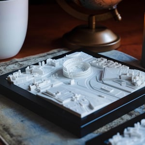 Puede incluir: Un modelo impreso en 3D blanco de una ciudad con un gran estadio en el centro, presentado en un marco negro. El modelo est&aacute; sobre una mesa con una vela y una copa de vino en el fondo.
