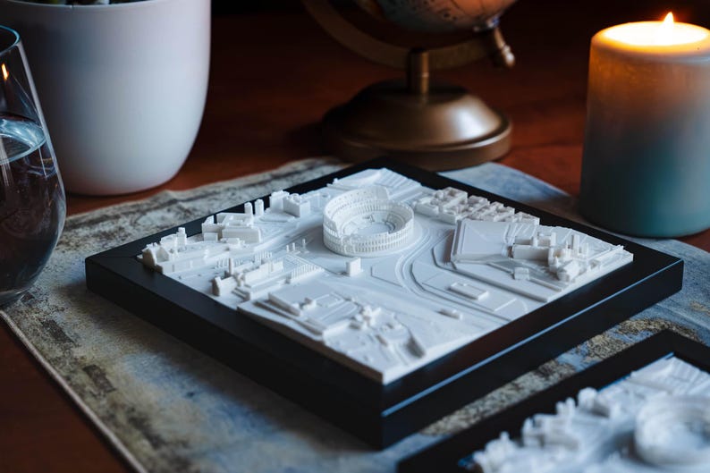 Puede incluir: Un modelo impreso en 3D blanco de una ciudad con un gran estadio en el centro, presentado en un marco negro. El modelo est&aacute; sobre una mesa con una vela y una copa de vino en el fondo.