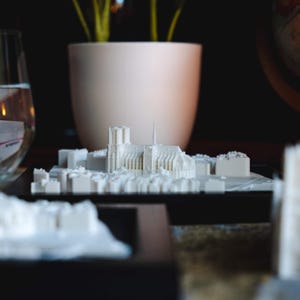 以下が含まれることがあります： 白い3Dプリントされた都市の街並みのモデルで、尖塔のある目立つ大聖堂が特徴です。モデルは黒い台座に置かれ、水が入ったグラスと鉢植えの植物のあるテーブルの上に置かれています。