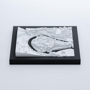 以下が含まれることがあります： 川が蛇行して流れる街の白い3Dモデル。モデルは黒い台座の上に置かれています。