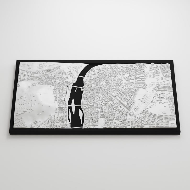 May include: A detailed, three-dimensional white map of a city, set within a black rectangular frame. The map features a prominent black river and bridge, with intricate building and street details. The overall aesthetic is modern and minimalist.