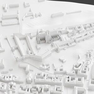 May include: A detailed, all-white architectural model of a city, showcasing various building types, including a cathedral with spires and multiple rectangular structures. The model is set against a white backdrop, emphasizing the intricate details of the urban landscape.