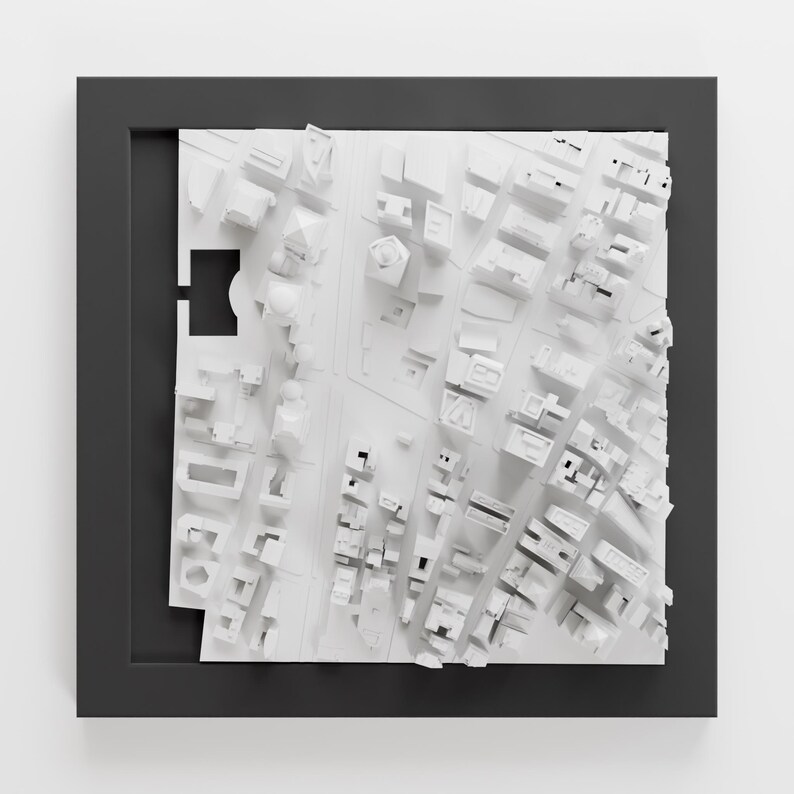 May include: A white 3D model of a city skyline, framed in a black frame. The model is made of paper or cardboard and shows a detailed cityscape with buildings, streets, and parks.