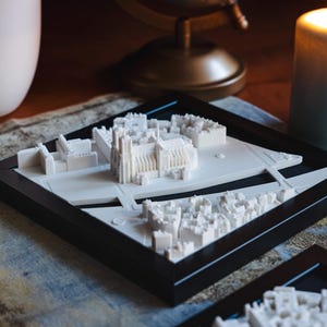 以下が含まれることがあります： 白い3Dプリントされた都市のモデルで、中央に大きな大聖堂があります。黒いフレームに収められ、質感のある表面に置かれています。