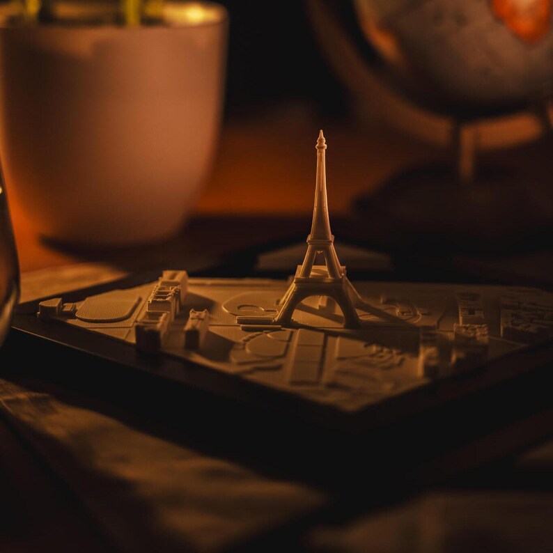 K&ouml;nnte beinhalten: Ein wei&szlig;es Plastikmodell des Eiffelturms auf einem wei&szlig;en Sockel mit einer Karte von Paris. Das Modell wird seitlich beleuchtet und wirft einen Schatten auf den Sockel.