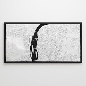 May include: A framed, three-dimensional white map with a black river running through it. The map is highly detailed, showing streets and buildings. The frame is black, and the overall aesthetic is modern and minimalist.