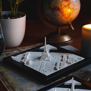 K&ouml;nnte beinhalten: Zwei schwarz gerahmte 3D-gedruckte Modelle des Eiffelturms und der umliegenden Pariser Stadtlandschaft. Die Modelle stehen auf einem Holztisch mit einem Globus und einer Kerze im Hintergrund.