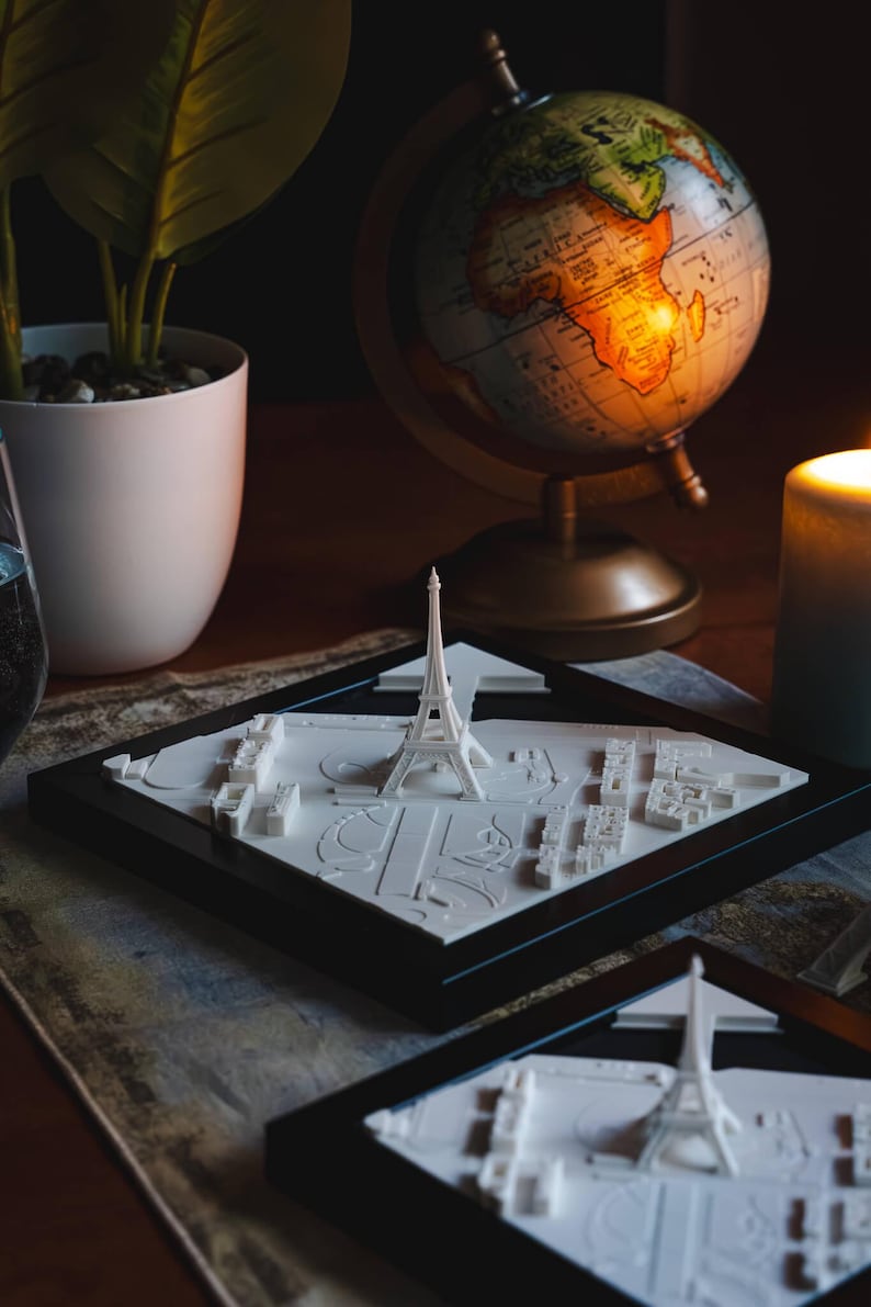 K&ouml;nnte beinhalten: Zwei schwarz gerahmte 3D-gedruckte Modelle des Eiffelturms und der umliegenden Pariser Stadtlandschaft. Die Modelle stehen auf einem Holztisch mit einem Globus und einer Kerze im Hintergrund.