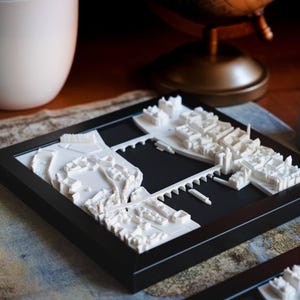 Puede incluir: Un modelo impreso en 3D de un horizonte de ciudad, con edificios blancos y una extensi&oacute;n de agua negra, enmarcado en un marco negro.