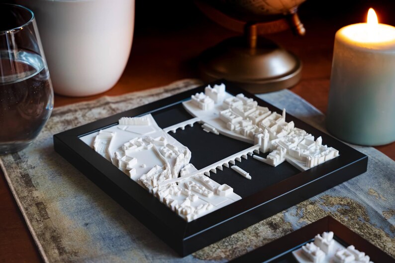 Puede incluir: Un modelo impreso en 3D de un horizonte de ciudad, con edificios blancos y una extensi&oacute;n de agua negra, enmarcado en un marco negro.