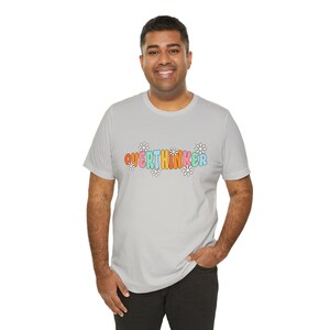 May include: A light gray t-shirt with the word "OVERTHINKER" in colorful, retro-style lettering. The text is surrounded by small white daisy flower graphics. The shirt is a casual, everyday wear item.
