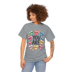 Könnte beinhalten: Ein graues T-Shirt mit einer bunten Grafik aus Bonbonherzen, auf denen "You Are" steht, umgeben von Wörtern wie "Strong", "Loved", "Brave", "Enough", "Capable" und "Worthy".
