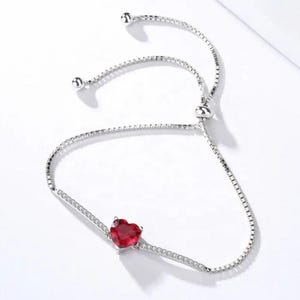 May include: A silver-toned adjustable bracelet featuring a central heart-shaped red gemstone. The band is composed of a delicate box chain with a sliding clasp mechanism and small clear stones embedded along the sides of the central heart setting.
