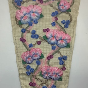 May include: A cream-colored textile with an embroidered floral design. Pink and blue pansies are arranged along a curved, ribbon-like pattern, accented with small blue and purple flowers and silver-colored beads.