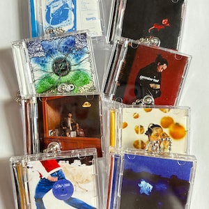 May include: A collection of eight miniature clear plastic CD case keychains, each featuring unique album cover artwork. The designs include various photographic and graphic prints, with metal ball chains attached to the top of each individual case.