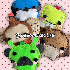 Puede incluir: Una colección de cinco llaveros de peluche de fieltro hechos a mano con varios diseños de personajes, incluyendo un pájaro azul, una esponja verde, un mono marrón, un oso marrón y un alienígena verde, con ojos de cuentas negras y cadenas.