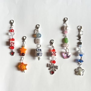 Könnte beinhalten: Eine Sammlung von Perlenanhängern mit verschiedenen Designs. Zu den Anhängern gehören rot-weiß gemusterte Perlen, sternförmige Perlen, eine Schleife mit einem Herz und eine Blume. Jeder Anhänger ist an einem silberfarbenen Verschluss befestigt.