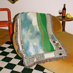 May include: A rectangular throw blanket with a pixelated landscape design in shades of blue, green, and gray. The blanket features a fringe trim and a graphic resembling a computer interface. The blanket is draped over a yellow armchair.