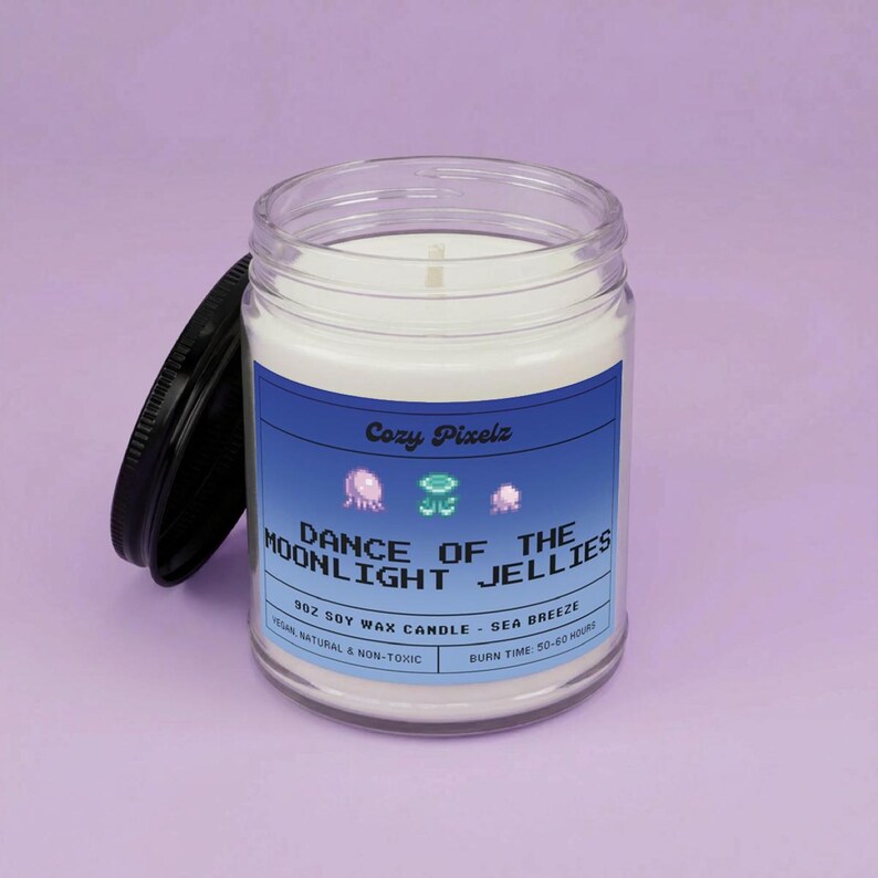 May include: A clear glass jar candle with a black lid, labeled "Dance of the Moonlight Jellies." The label is blue with pixelated jellyfish graphics. The candle is filled with white wax and has a burn time of 50-60 hours.