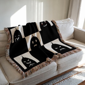 Puede incluir: Manta de tejido negro y blanco con un patrón de cuadros que presenta un diseño repetido de caras de calabazas negras. La manta tiene flecos alrededor de los bordes.