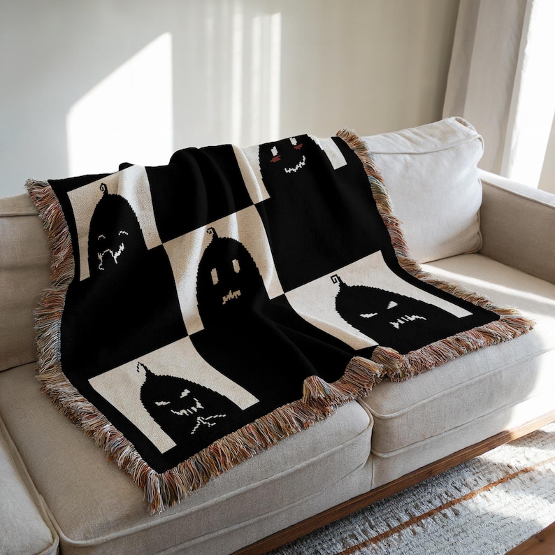 May include: Black and white woven throw blanket with a checkered pattern featuring a repeated design of black jack-o'-lantern faces. The blanket has fringe around the edges.